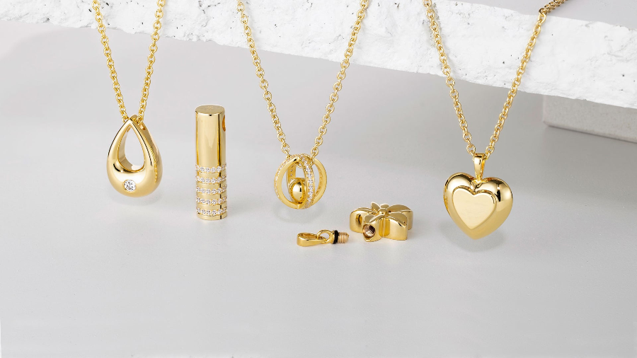 A jewelry set in polished yellow gold, featuring four pendant necklaces (teardrop, cylinder, geometric, heart), a matching cylindrical box, and a pair of stud earrings, arranged on a light gray surface with a white textured block.
