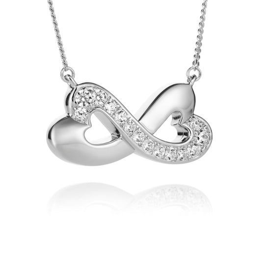 A silver infinity-shaped pendant necklace featuring two interlocking hearts, one studded with small clear crystals, suspended from a delicate silver chain against a white background with a soft reflection below.