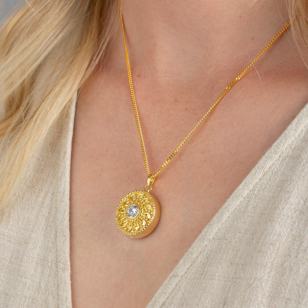 A close-up of a model wearing a gold pendant necklace with a textured circular medallion and a clear central gemstone, styled with a beige linen top.