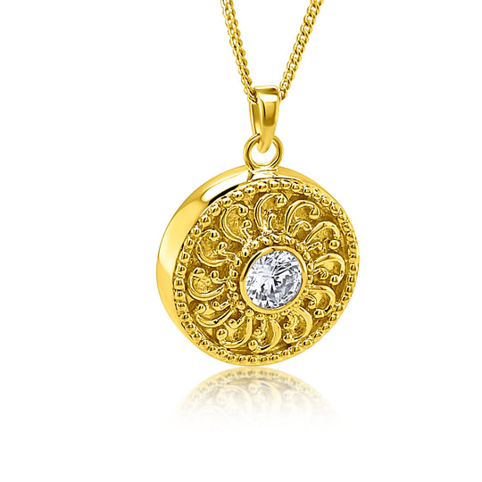 A gold round pendant necklace with intricate filigree detailing and a central clear gemstone, shown on a white background with a soft reflection.