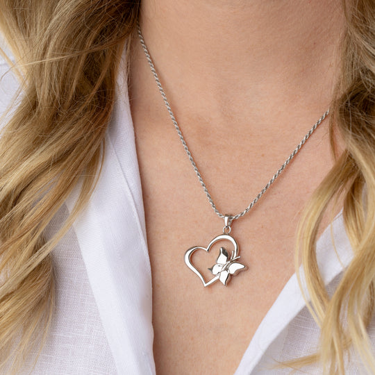 A close-up of a woman wearing a silver necklace with a heart-shaped pendant and a small butterfly detail, styled with a white collared shirt.