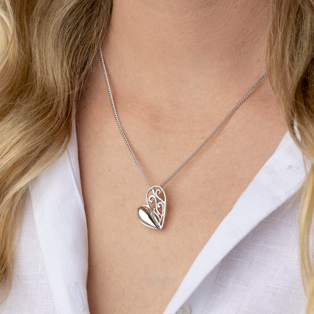 A close-up of a woman wearing a delicate silver necklace with a heart-shaped pendant. The pendant features an openwork floral design on one side and a smooth, polished heart on the other.