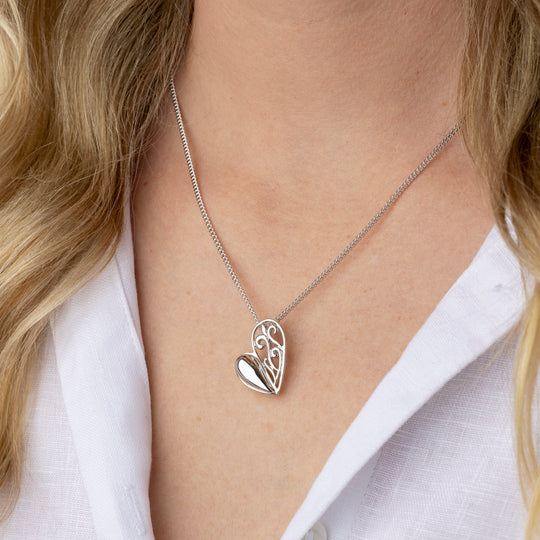 A close-up of a woman wearing a delicate silver necklace with a heart-shaped pendant. The pendant features an openwork floral design on one side and a smooth, polished heart on the other.