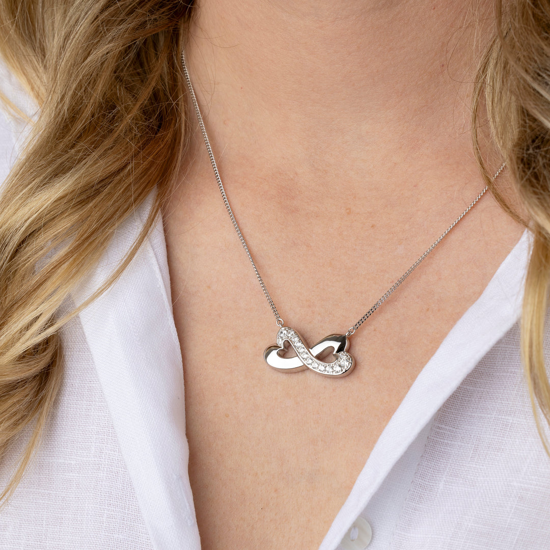 A close-up of a woman wearing a delicate silver infinity necklace with a small crystal accent, styled with a white linen shirt.