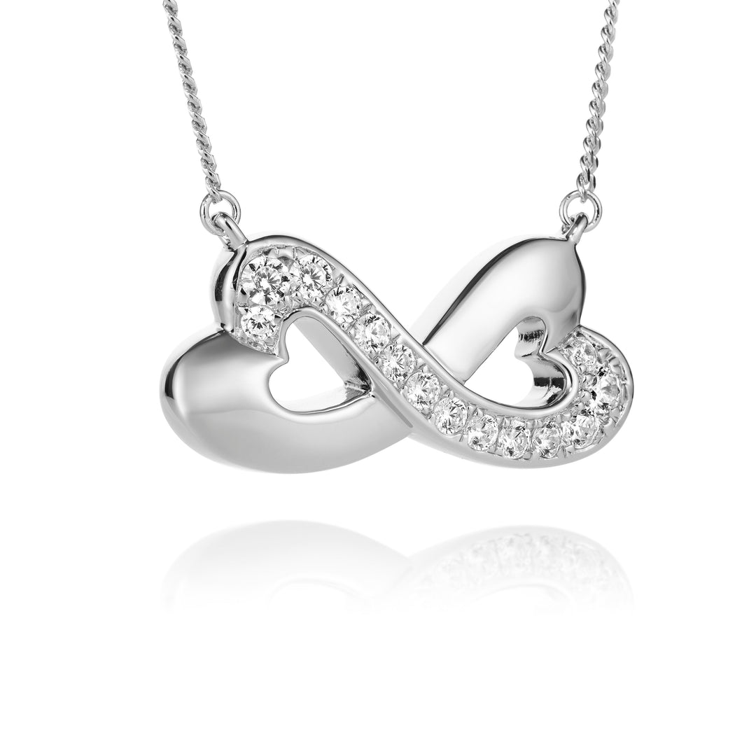 A silver infinity-shaped pendant necklace featuring two interlocking hearts, one studded with small clear crystals, suspended from a delicate silver chain against a white background with a soft reflection below.