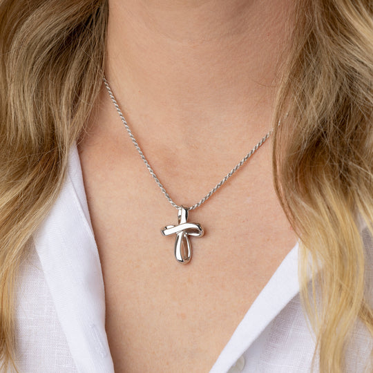 A close-up of a person wearing a delicate silver cross pendant necklace on a fine chain, styled with a white collared shirt.