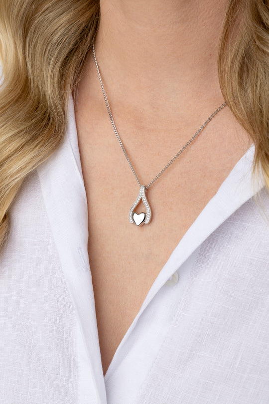 A close-up of a woman wearing a delicate silver chain necklace with a teardrop-shaped pendant featuring a small heart in the center, styled with a white collared shirt.