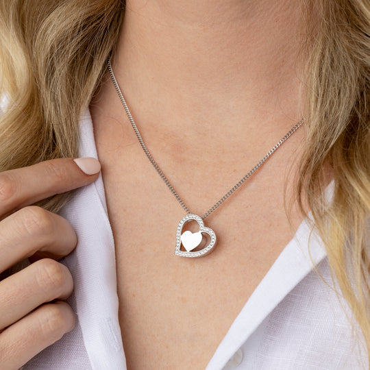 A close-up of a model wearing a silver heart pendant necklace with a smaller inner heart, styled with a white linen shirt.