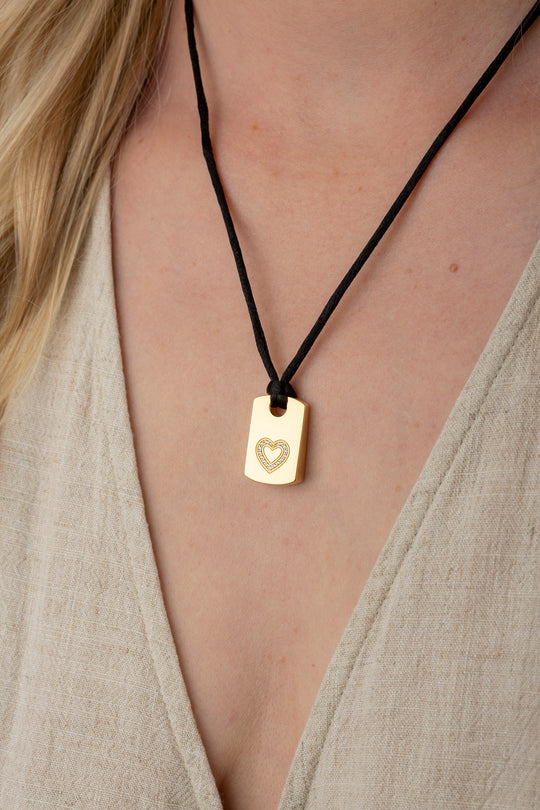 A close-up of a person wearing a gold rectangular pendant necklace with a small heart design on a black cord, paired with a beige linen top.