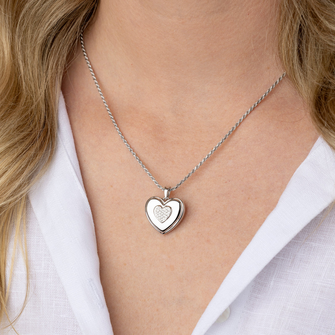 A silver heart-shaped pendant necklace worn by a model in a white collared shirt. The pendant features a polished outer heart and a smaller, textured heart design inside.