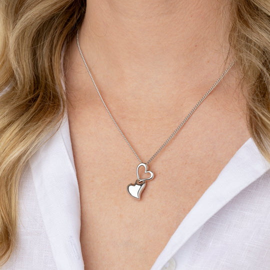 A close-up of a model wearing a delicate silver chain necklace with two interlocking heart pendants, styled with a white collared shirt.