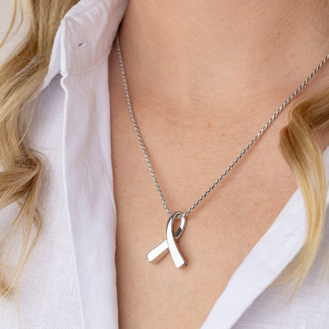 A close-up of a woman wearing a silver awareness ribbon pendant necklace on a delicate silver chain, styled with a white collared shirt.