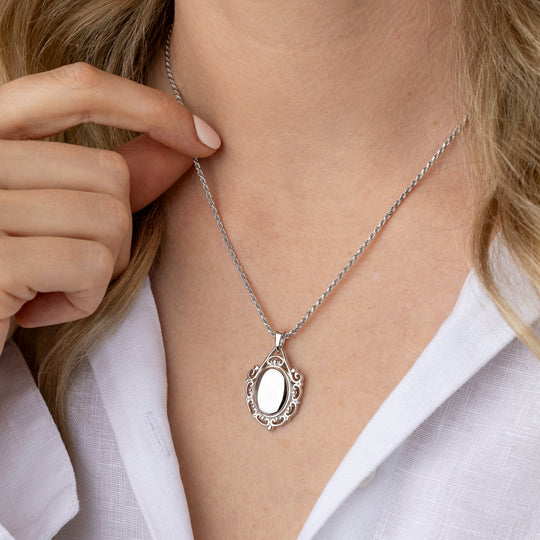 A close-up of a woman wearing a silver oval pendant necklace with a decorative filigree border and a shiny inlay, styled with a white collared shirt.