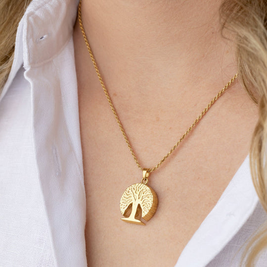 A gold necklace with a circular pendant featuring a tree of life design, worn by a model in a white collared shirt.