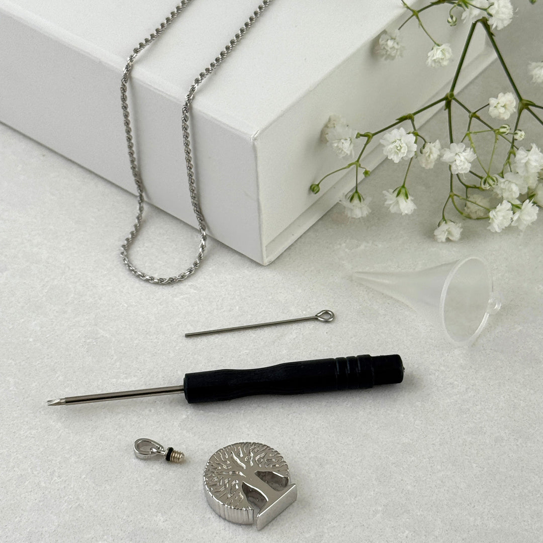 Silver chain necklace with a tree-of-life pendant displayed next to its white Ashgate brand box, along with a black-handled tool, small metal parts, and baby&